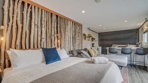 A modern bedroom with a large bed, a wooden headboard, and a kitchenette in the background. - PeakView Lil Lux! Hot Tub & Mountain Views (Colorado Springs)