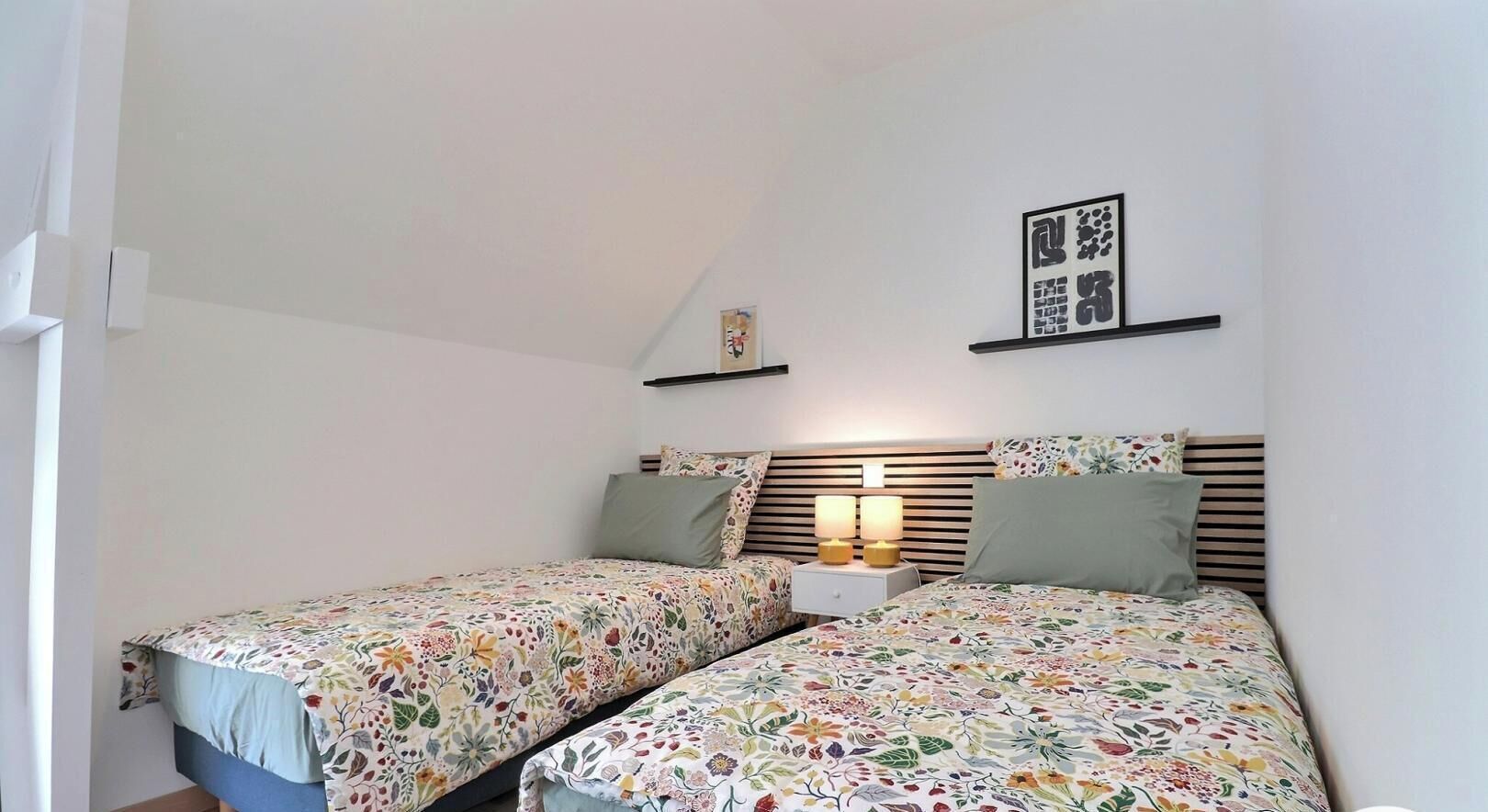A bedroom with two beds, a nightstand with lamps, a shelf with framed pictures, and a sloped ceiling.