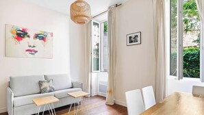 A living room with a grey sofa, two small tables, a hanging light fixture, a framed picture, and a window with curtains. - Suite Lumineuse en plein coeur du Marais (Paris)