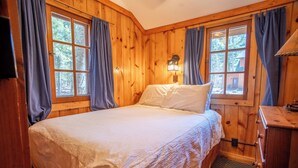 Ein Schlafzimmer einer Holzhütte mit Bett, Vorhängen, einem Nachttisch und einem Fenster.