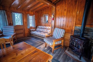 A cozy cabin interior with wooden walls, a sofa, two plaid chairs, a wooden table, and a wood-burning stove. - The Fox Cabin at Timber Bay (Babbitt)
