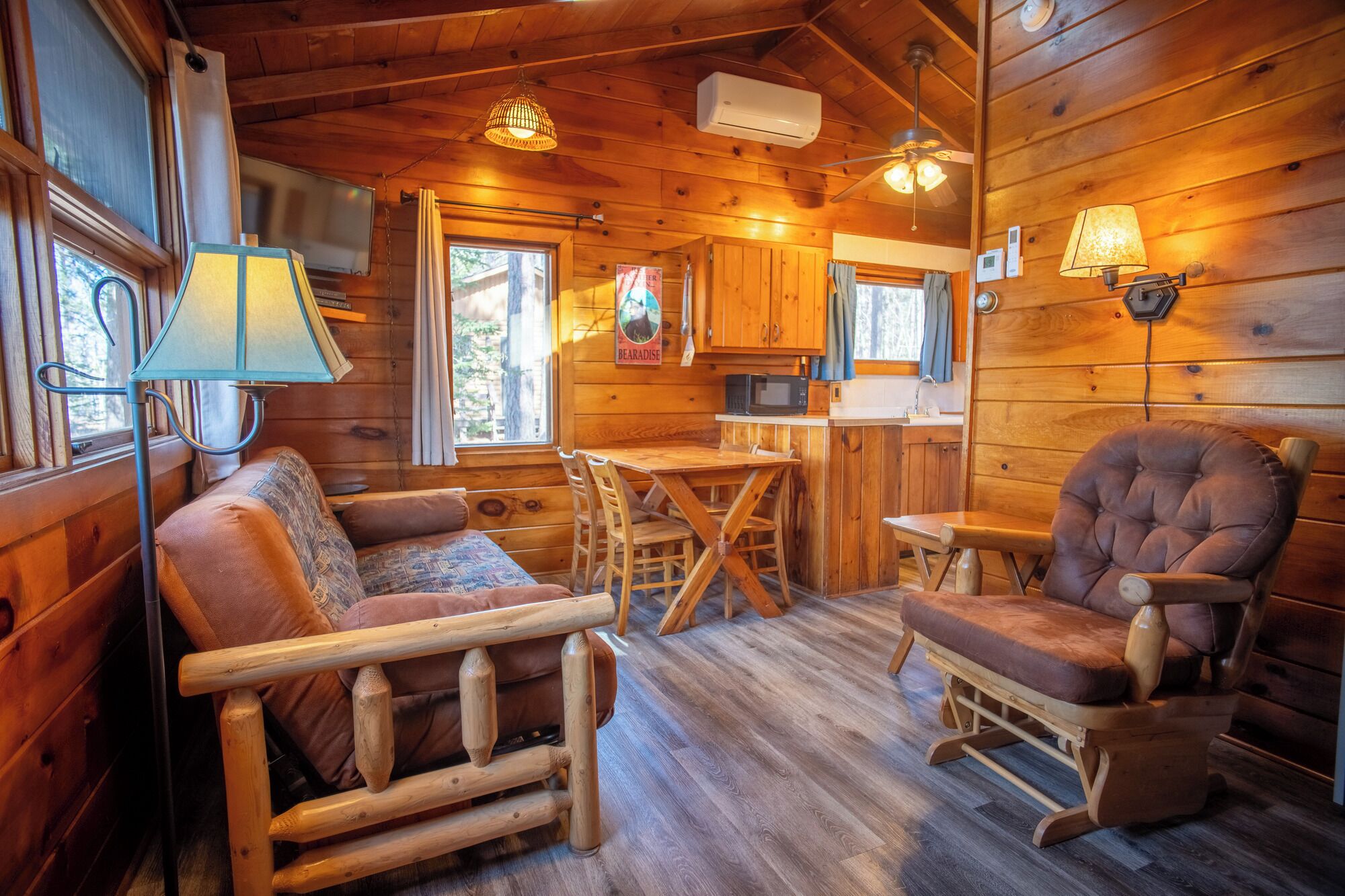 A cozy cabin interior with a wooden dining table, two cushioned chairs, a kitchen area, and a window with curtains.