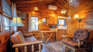 A cozy cabin interior with a wooden dining table, two cushioned chairs, a kitchen area, and a window with curtains. - For our honeymooners: The Hummingbird Cabin (Babbitt)