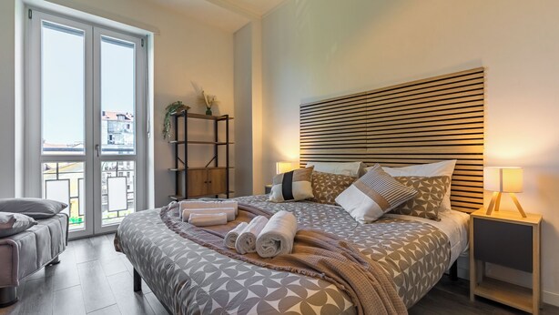 A modern bedroom with a large bed, a bedside table with a lamp, a view through the sliding doors, and a small seating area. - In Turin bright apartment, for those seeking comfort and tranquility (Torino)