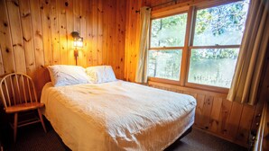 Habitación con paneles de madera, una cama, una silla y una ventana con vistas a árboles.