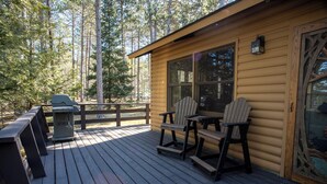 Una cabaña de madera con un porche, dos sillas Adirondack, una parrilla y una ventana con vistas a un bosque.