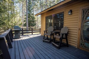 A wooden cabin with a deck, two Adirondack chairs, a grill, and a window overlooking a forest. - The Martin Cabin at Timber Bay (Babbitt)