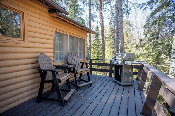 Eine Blockhütte mit einer Terrasse, zwei Adirondack-Stühlen und einem Grill.