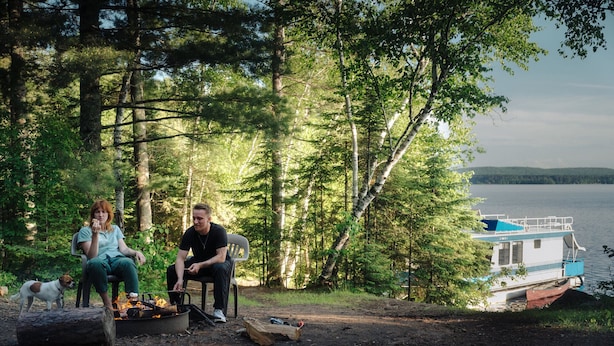 Two people sitting by a campfire in a forested area with a boat on the water in the background. - The Mallard Houseboat at Timber Bay (Babbitt)