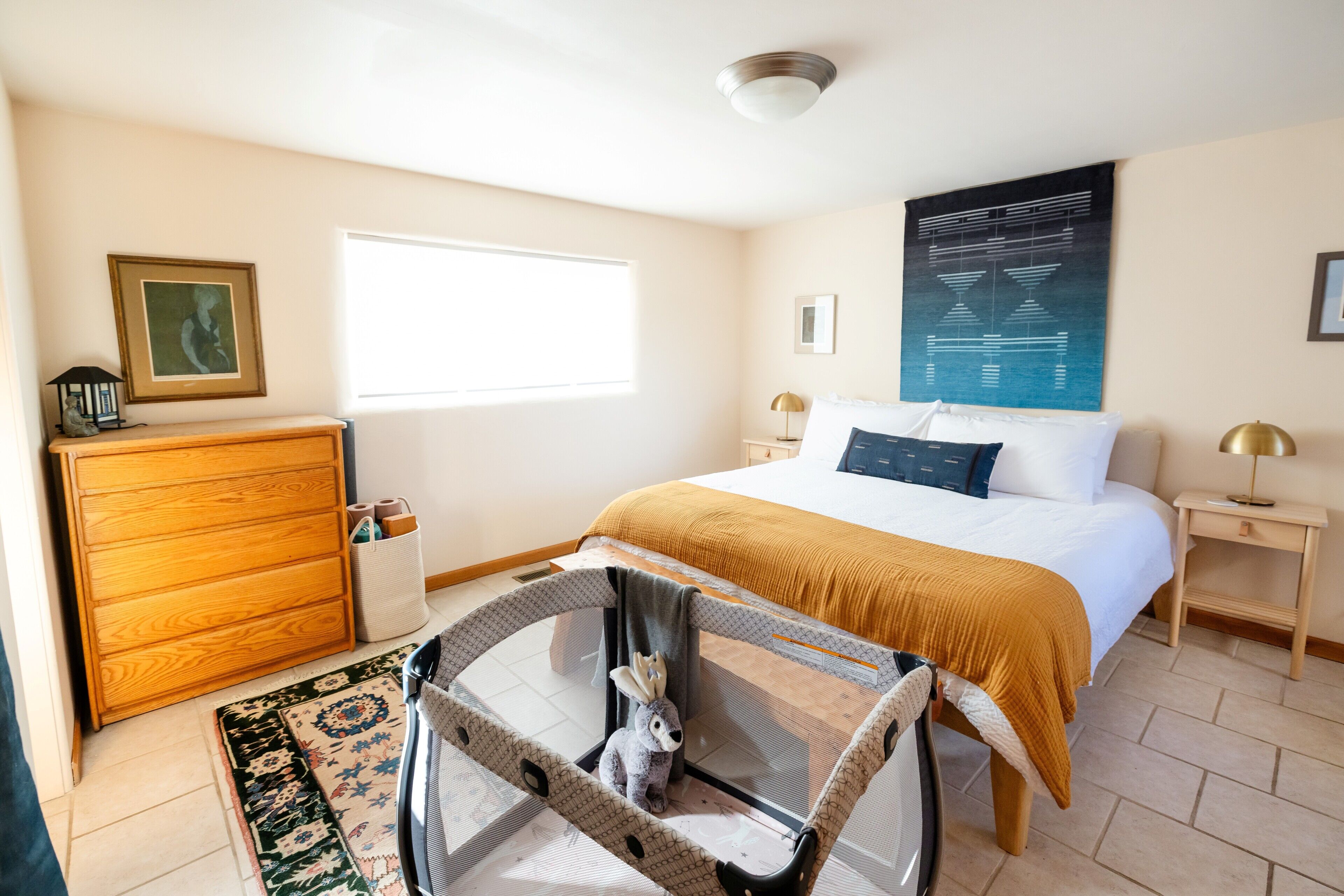 Un dormitorio con cama, cómoda de madera, mesita con silla, ventana y una obra de arte colgada en la pared.