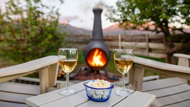 A wooden deck with two chairs, a bowl of popcorn, and two glasses of white wine, with a wood-burning stove in the background.