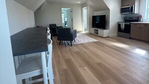 En rummelig opholdsstue med spisebord, stole, fjernsyn og et køkken med hårde hvidevarer.