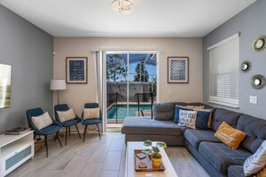 Ein Wohnzimmer mit einer Couch, Sesseln, einem Couchtisch und einem Fernseher. Ein Glastür führt zu einem Pool.