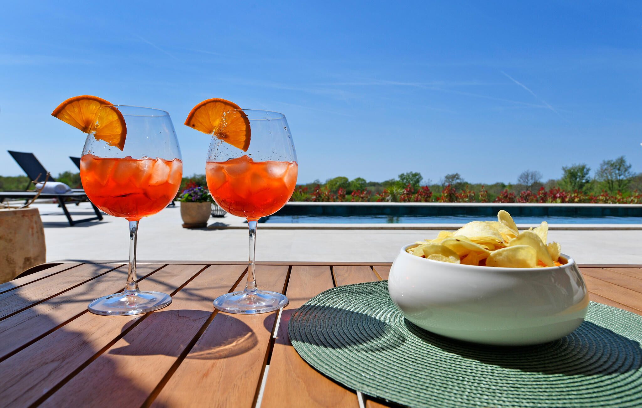 Two glasses of red drink with orange slices on a wooden table, a bowl of chips, and a pool in the background.