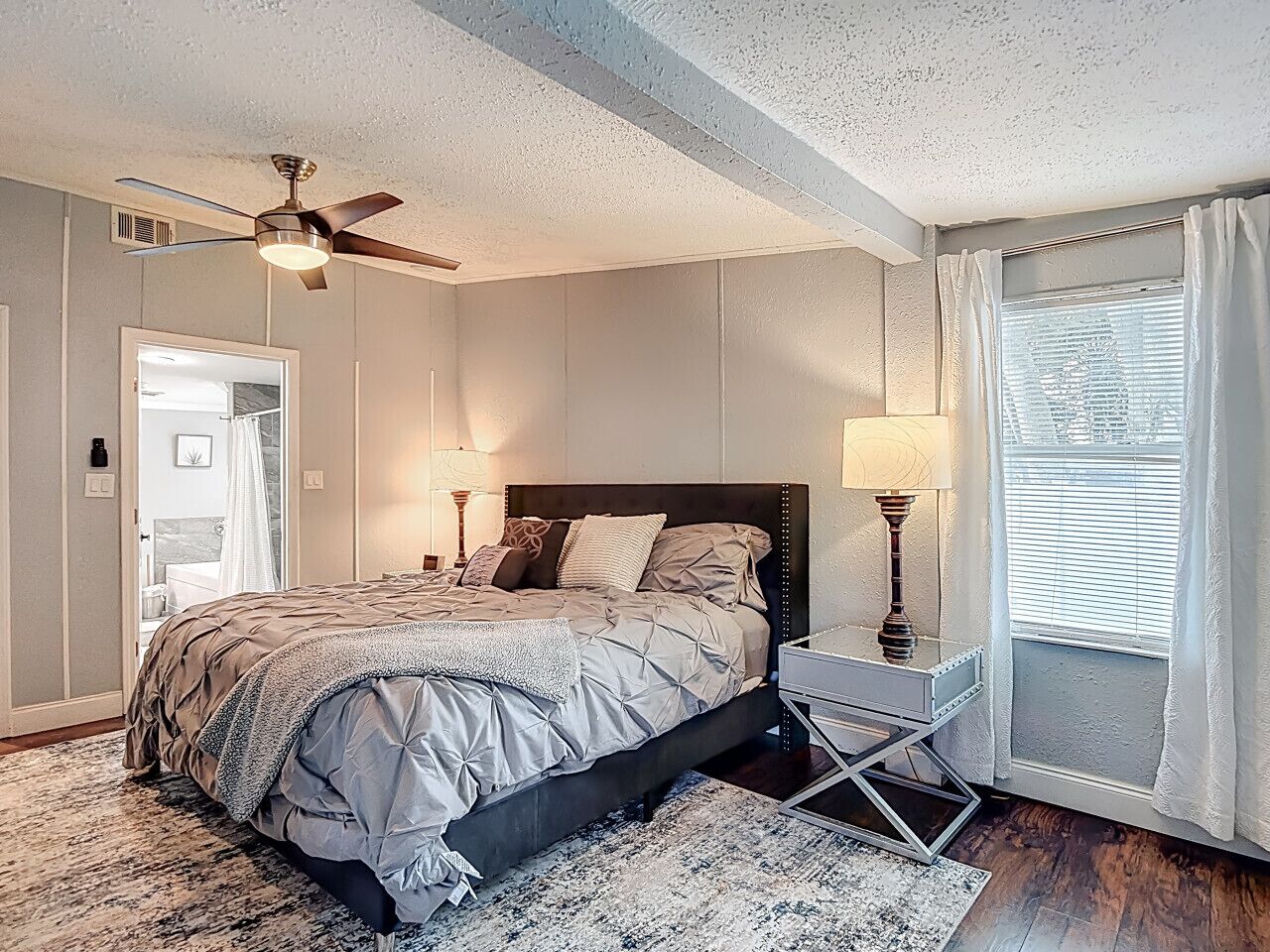 A bedroom with a bed, bedside tables, a ceiling fan, a window with curtains, and a door leading to another room.