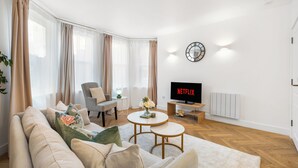 A modern living room with a sofa, armchairs, a coffee table, a TV, and a clock on the wall. - Stylish Seaside Apartment | Minutes from Beach (Eastbourne)