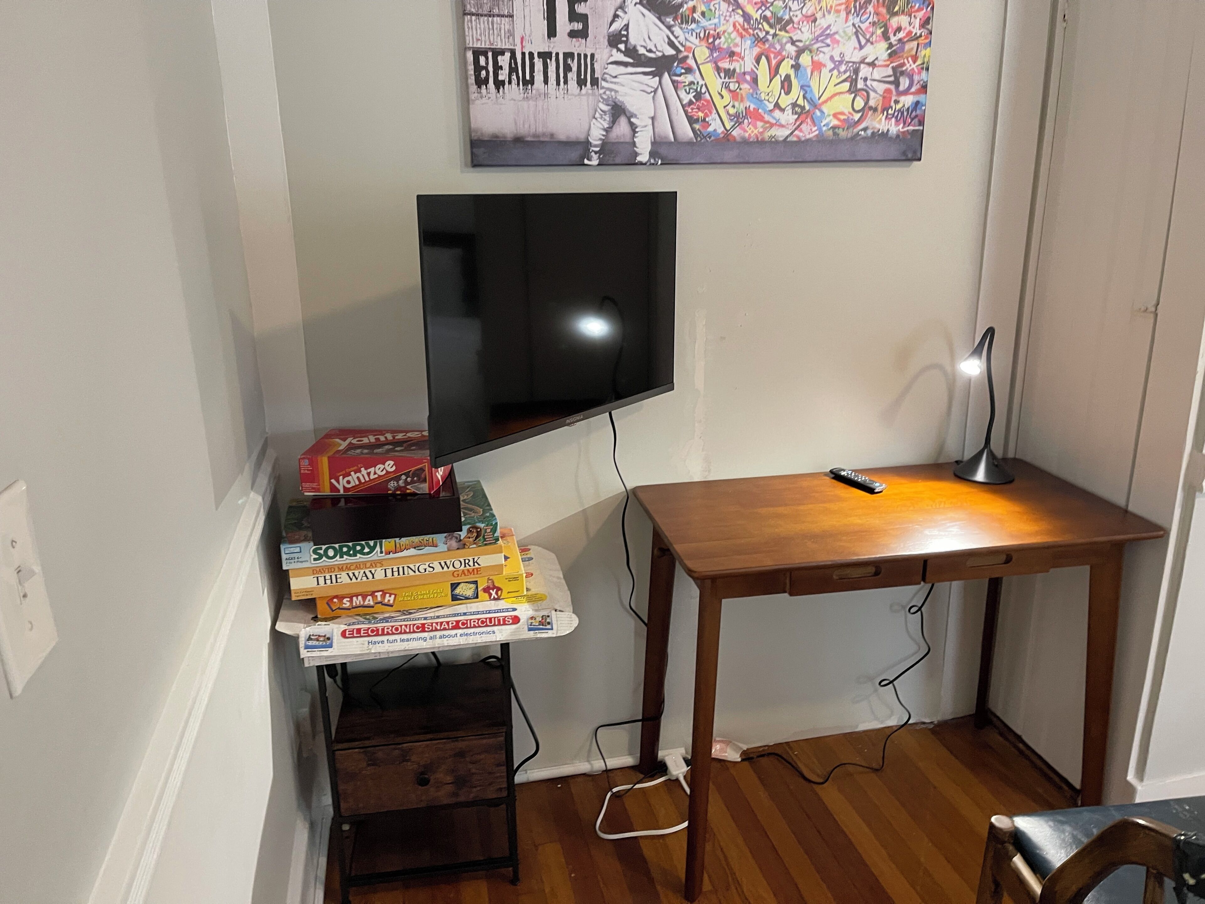 A room with a television mounted on the wall, a wooden desk with a lamp, and a shelf with electronic games.