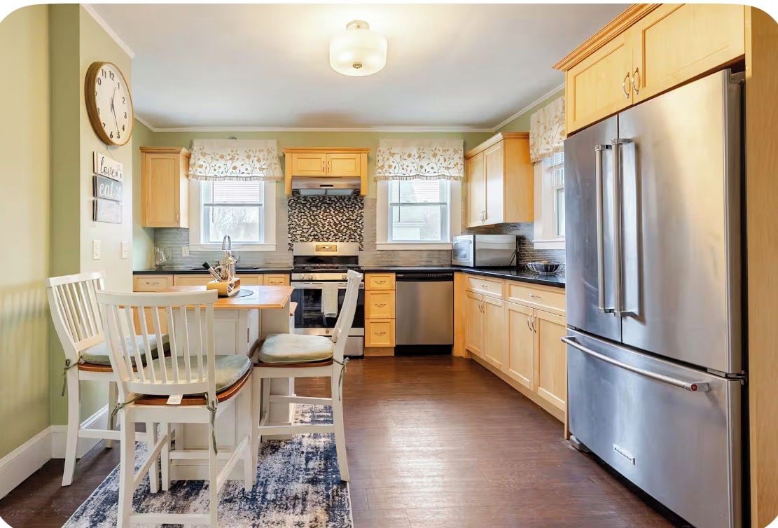 A kitchen with wooden cabinets, a stainless steel refrigerator, a dining table with chairs, and a clock on the wall.