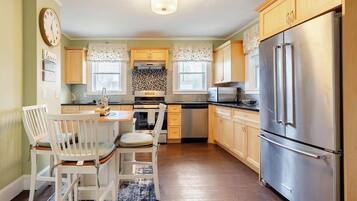 A kitchen with wooden cabinets, a stainless steel refrigerator, a dining table with chairs, and a clock on the wall.