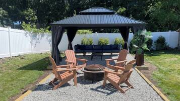 A gazebo with a fire pit and Adirondack chairs in a backyard.