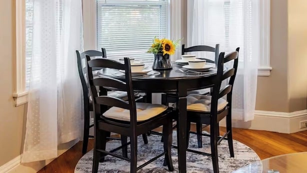 A dining area with a round table, four chairs, a vase with flowers, and sheer curtains. - 4BR Family Vacation Home • Near Boston • Gillette Stadium   • Free Parking (Norwood)