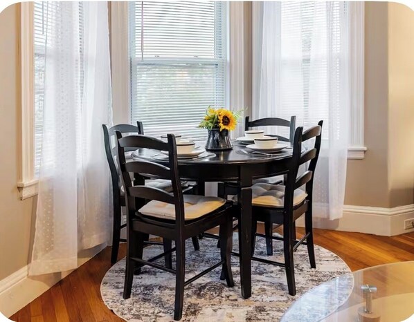 A dining area with a round table, four chairs, a vase with flowers, and sheer curtains. - 4BR Family Vacation Home • Near Boston • Gillette Stadium   • Free Parking (Norwood)