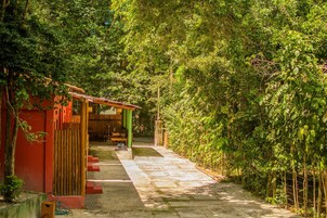 Uma trilha leva a um edifício com fachada vermelha e telhado verde, cercado por vegetação densa.