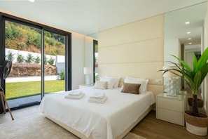 A modern bedroom with a large bed, bedside tables, a mirror, and a view of a garden. - 202 - Casinha da Ribeira (Vila Verde)