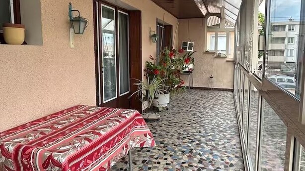 Ein überdachter Balkon mit Fliesenboden, einem Tisch mit gemustertem Tischläufer und Topfpflanzen.