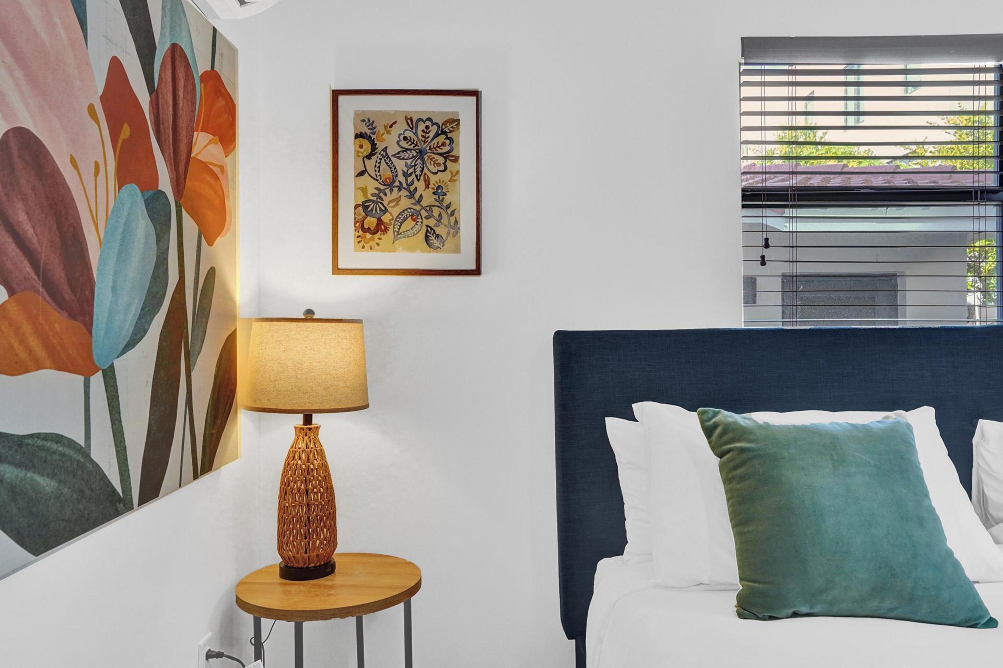 A bedroom with a bed, a lamp, a painting, and a window with blinds.