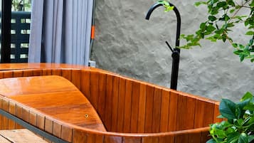 Una bañera de madera con un banco, una toalla y una botella sobre una mesa, frente a una pared texturizada con una planta y una estructura metálica curva.