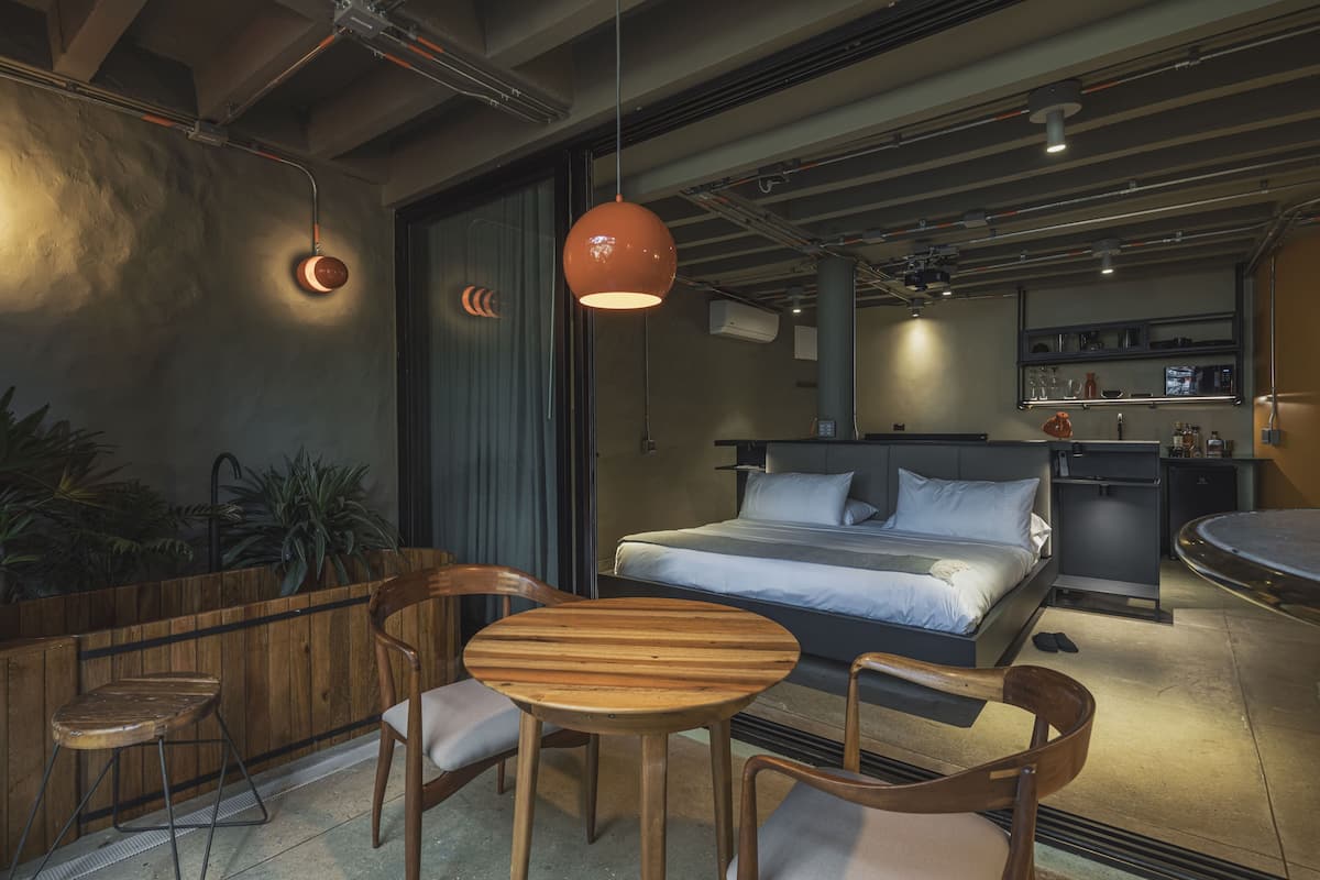 A modern hotel room with a bed, a round wooden table, two chairs, a small kitchen area, and a seating area with plants.