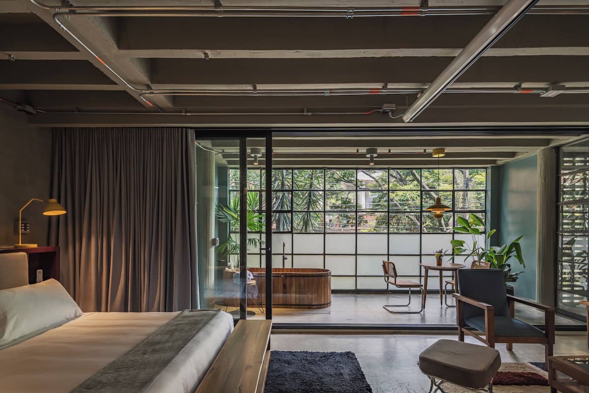 A modern hotel room with a large bed, a desk, a chair, a small table, a sofa, a bench, a bathtub, and a view of greenery through large windows.