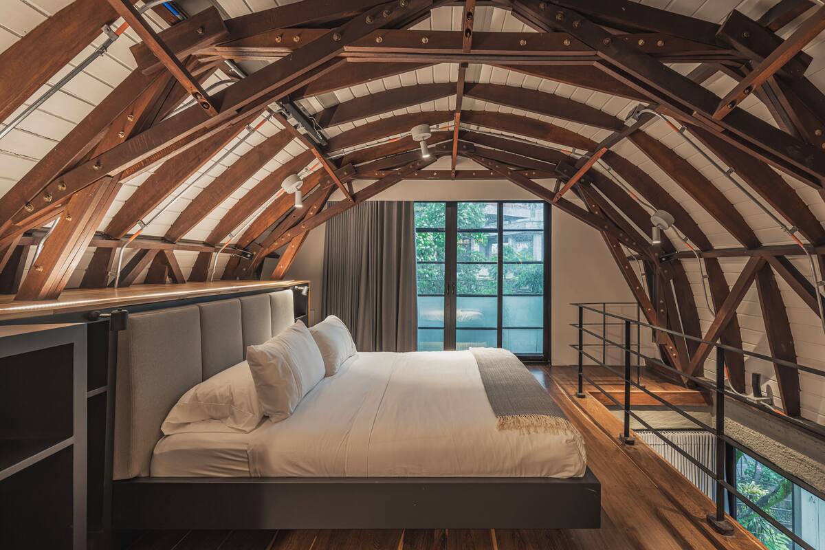 A high-ceilinged bedroom with a large bed, wooden beams, and a view of greenery outside.
