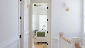 A hallway with white walls, wooden flooring, a staircase with white railings, and a door leading to a bedroom with a bed and a window. - Stunning Downtown 3BD-2.5BA Home| Near Forsyth Pk (Savannah)