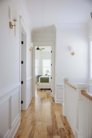 Ein Flur mit weißen Wänden, Holzdielen, einer Treppe mit weißen Geländern und einer Tür zu einem Schlafzimmer mit Bett und Fenster.