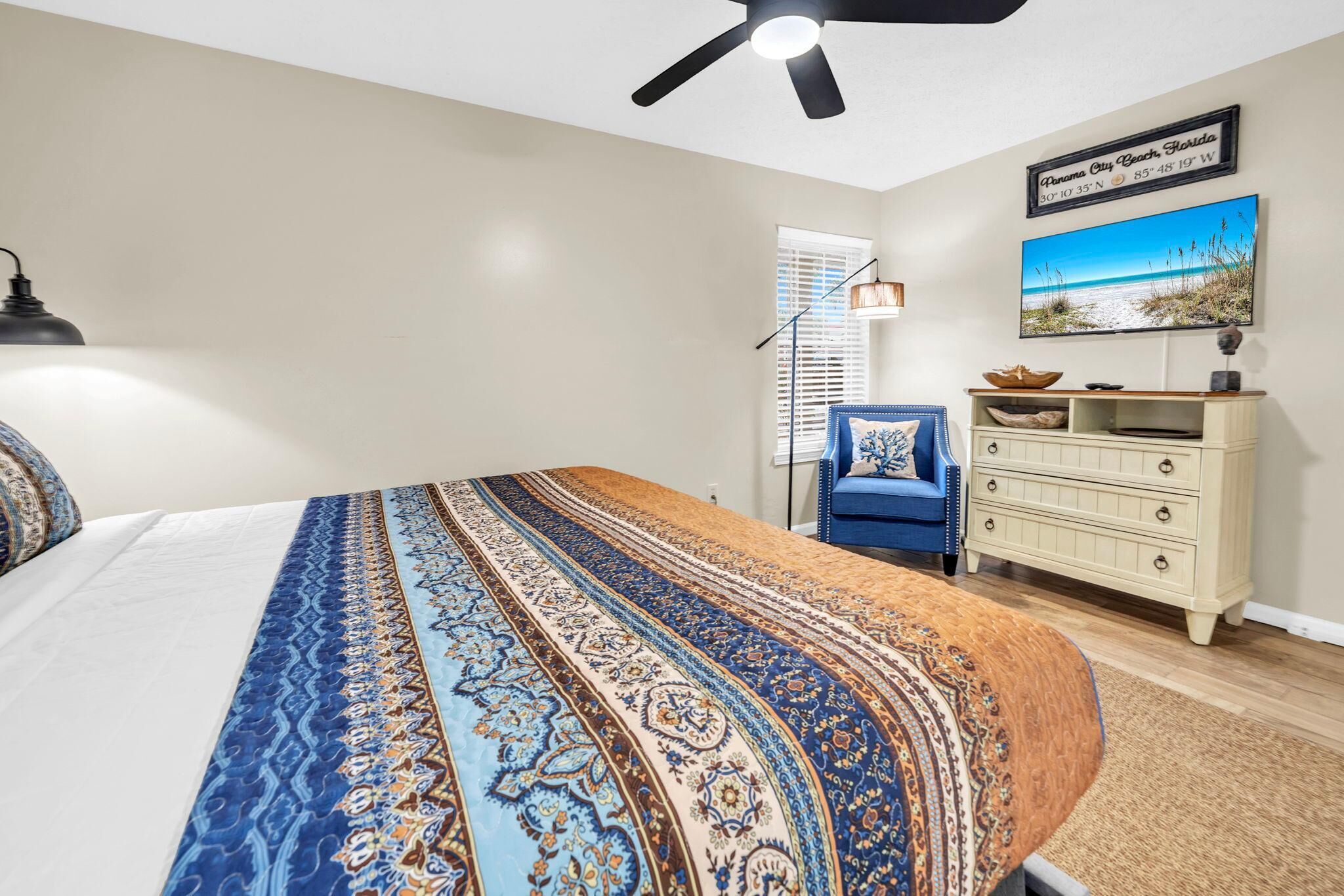 A bedroom with a bed, a ceiling fan, a dresser, a chair, and a wall-mounted TV.