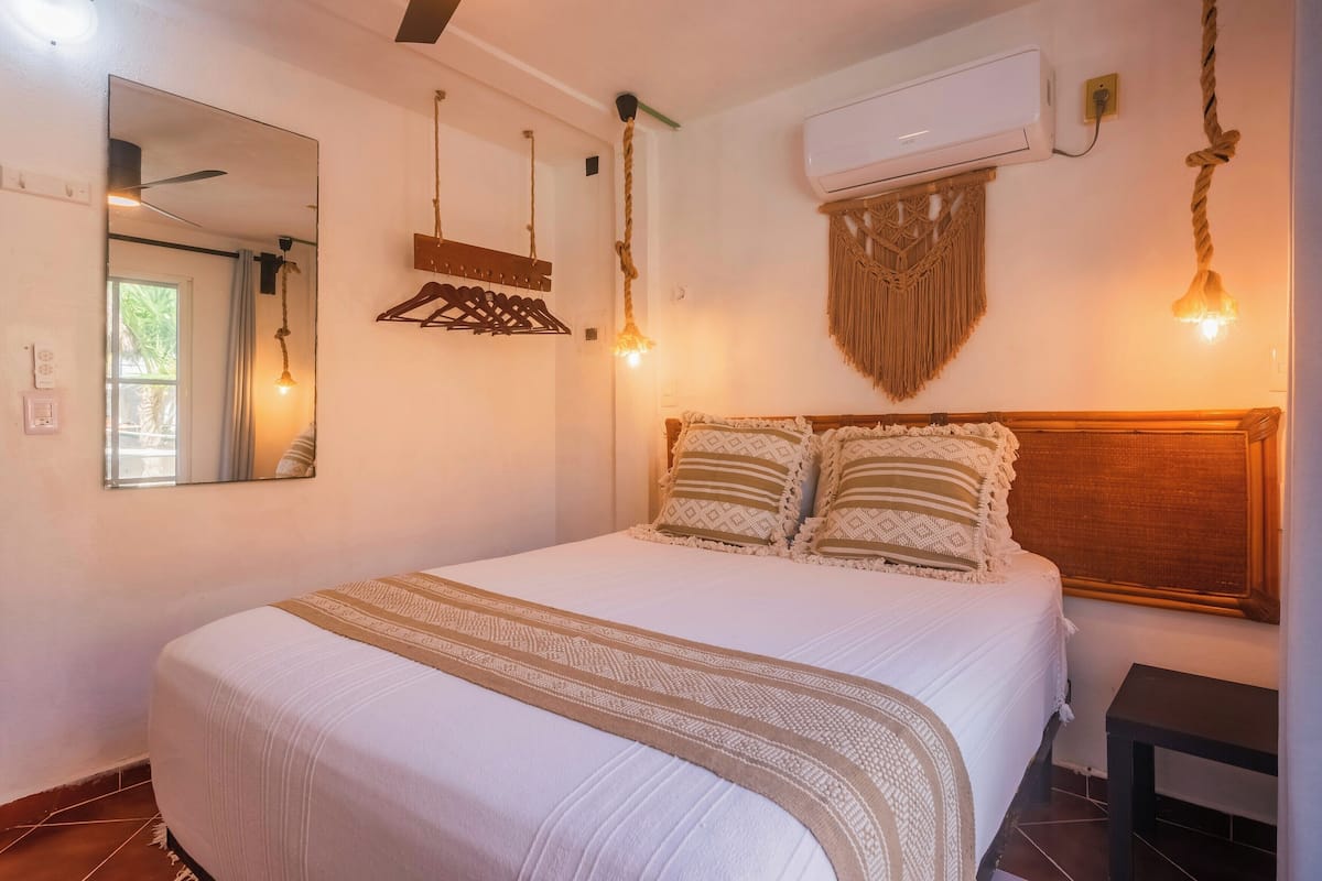 Una cama bien hecha con una colcha estampada, una lámpara colgante, un aire acondicionado instalado en la pared y un espejo.