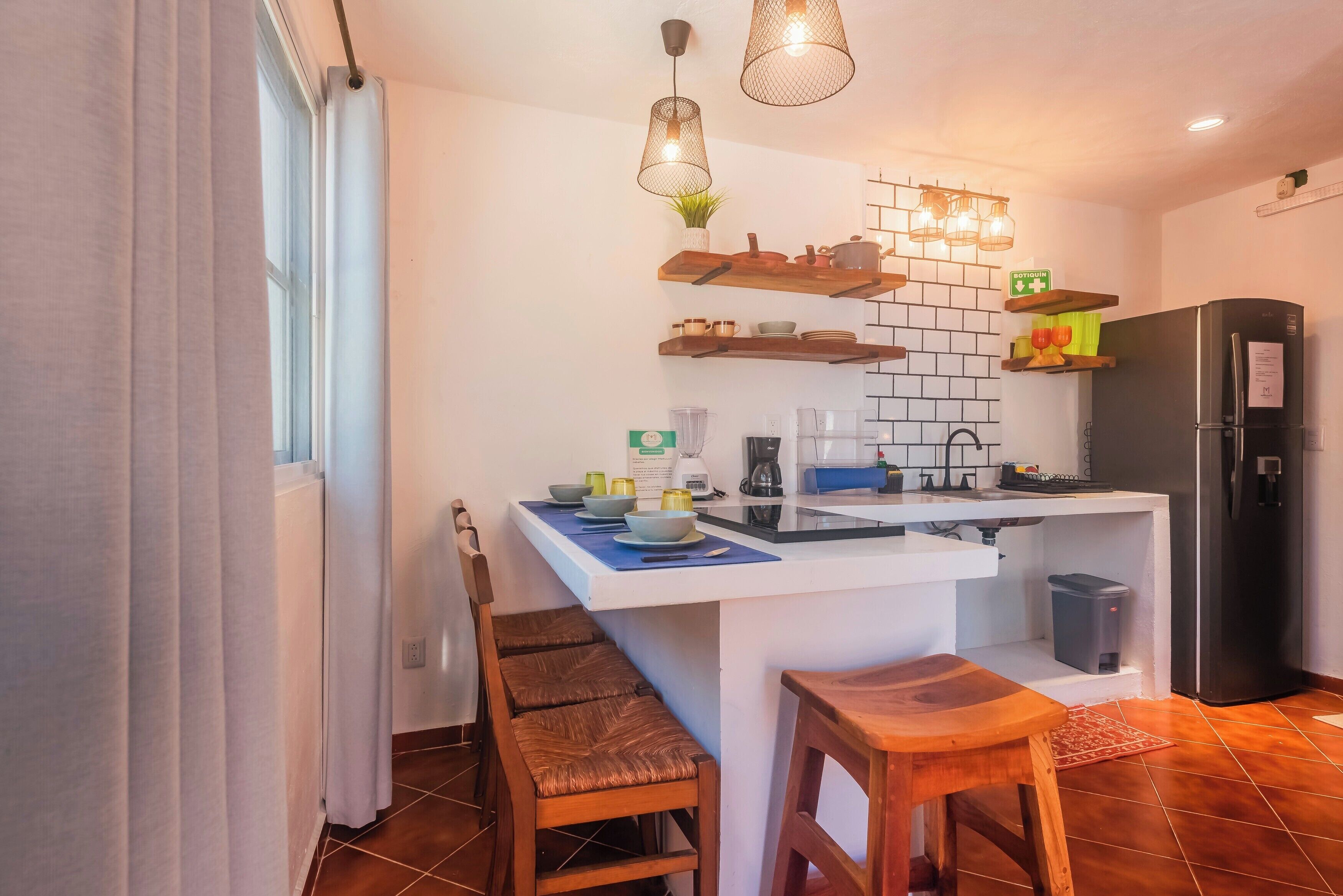 A compact kitchen with a white countertop, wooden chairs, a black refrigerator, and pendant lights.