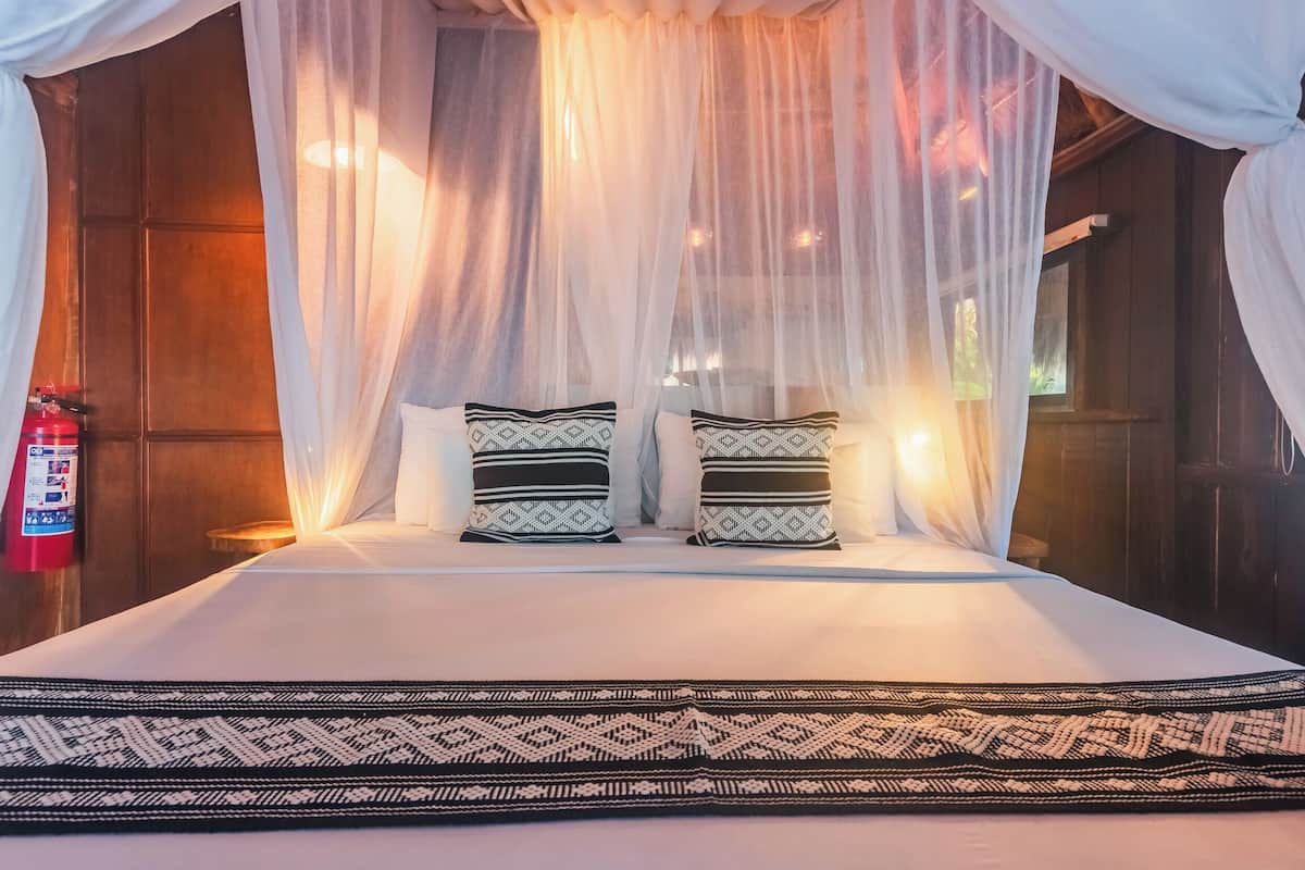 Una cama con dosel, dos almohadas estampadas, un tapete estampado y un extintor en la pared.