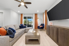 Ein modernes Wohnzimmer mit einem Ecksofa, einem Couchtisch, einem an der Wand montierten Fernseher und einem Deckenventilator.