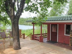 En träveranda med en röd stuga, ett träd och utsikt över en sjö.
