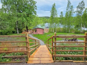 A wooden bridge leading to a red building with a green roof, surrounded by lush greenery and a body of water.