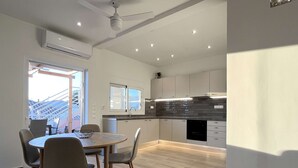 A modern kitchen with a dining area, wooden flooring, and a view through a large window. - Taygetos View - Bright Modern Retreat in Center (Kalamata)