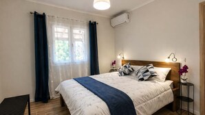 Une chambre à coucher comprenant un lit, une table de chevet, un banc, une fenêtre avec des rideaux, un appareil de climatisation et un luminaire fixé au mur.