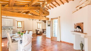 A spacious room with a high ceiling, exposed wooden beams, a fireplace, a dining area with a table set for two, and a cozy seating area. - Villa La Bergerie by Villa Plus (Saint-Cézaire-sur-Siagne)
