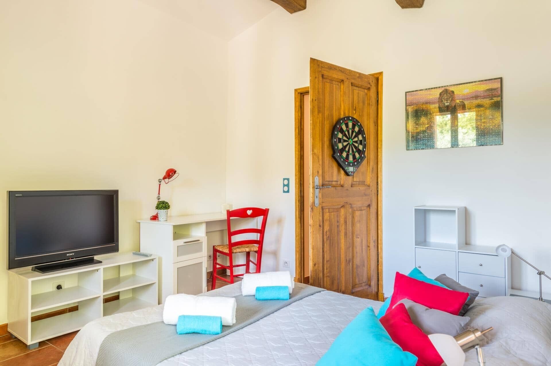 Un dormitorio con cama, televisor, puerta de madera, dardo y un cuadro.