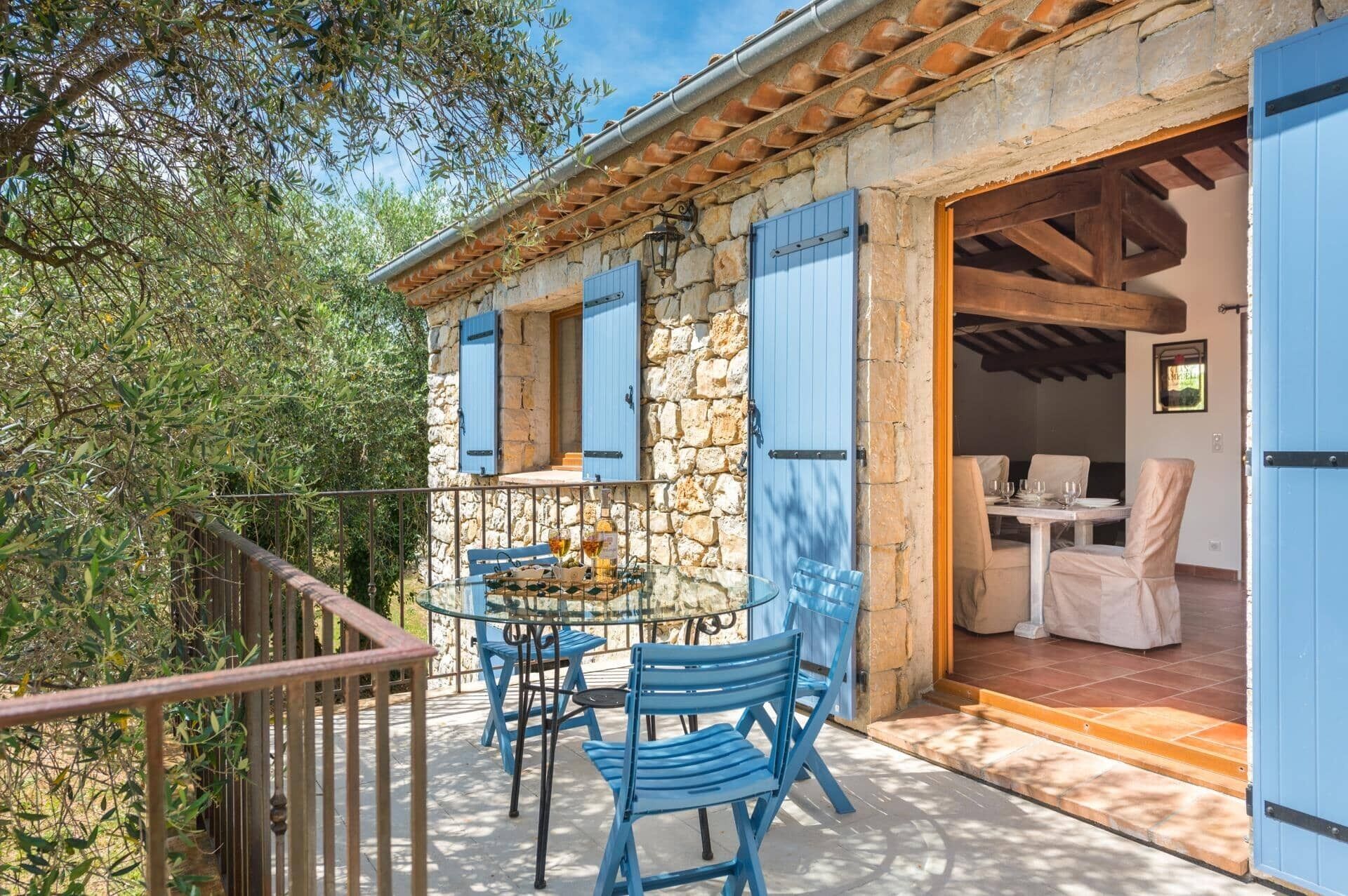 Un edificio de piedra con contraventanas azules, un balcón con sillas azules y una mesa con superficie de vidrio.