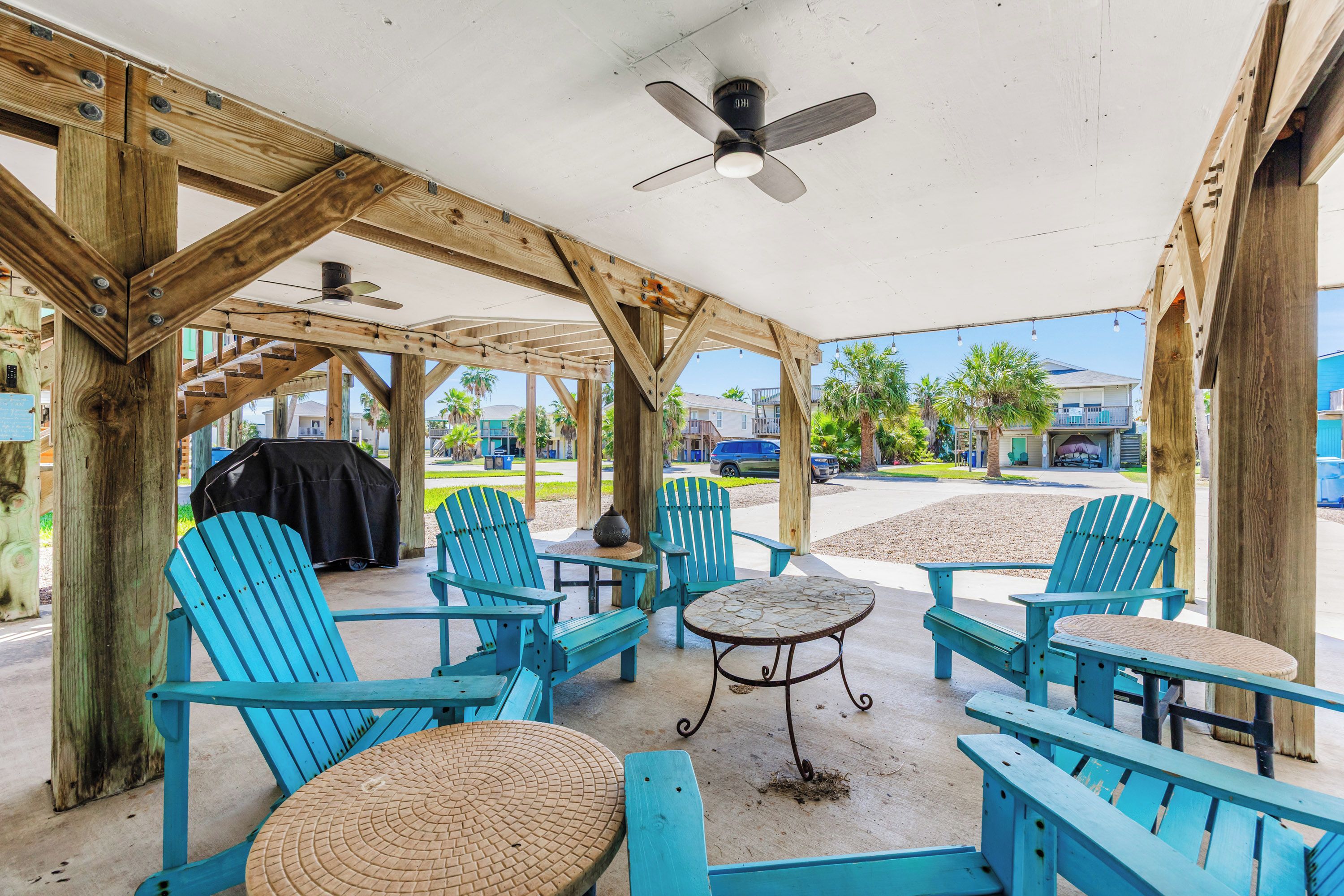 A covered outdoor seating area with blue Adirondack chairs and a small round table, under a wooden structure with a ceiling fan.