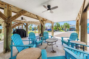 A covered outdoor seating area with blue Adirondack chairs and a small round table, under a wooden structure with a ceiling fan.